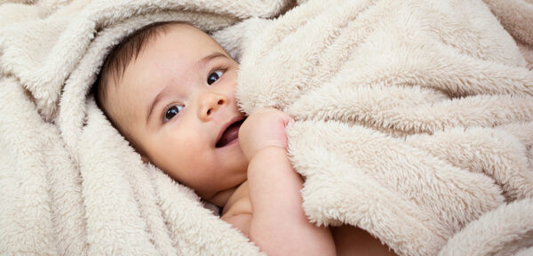 Ein Baby kuschelt sich in eine besonders flauschige, cremefarbene Decke und schaut neugierig in die Kamera.