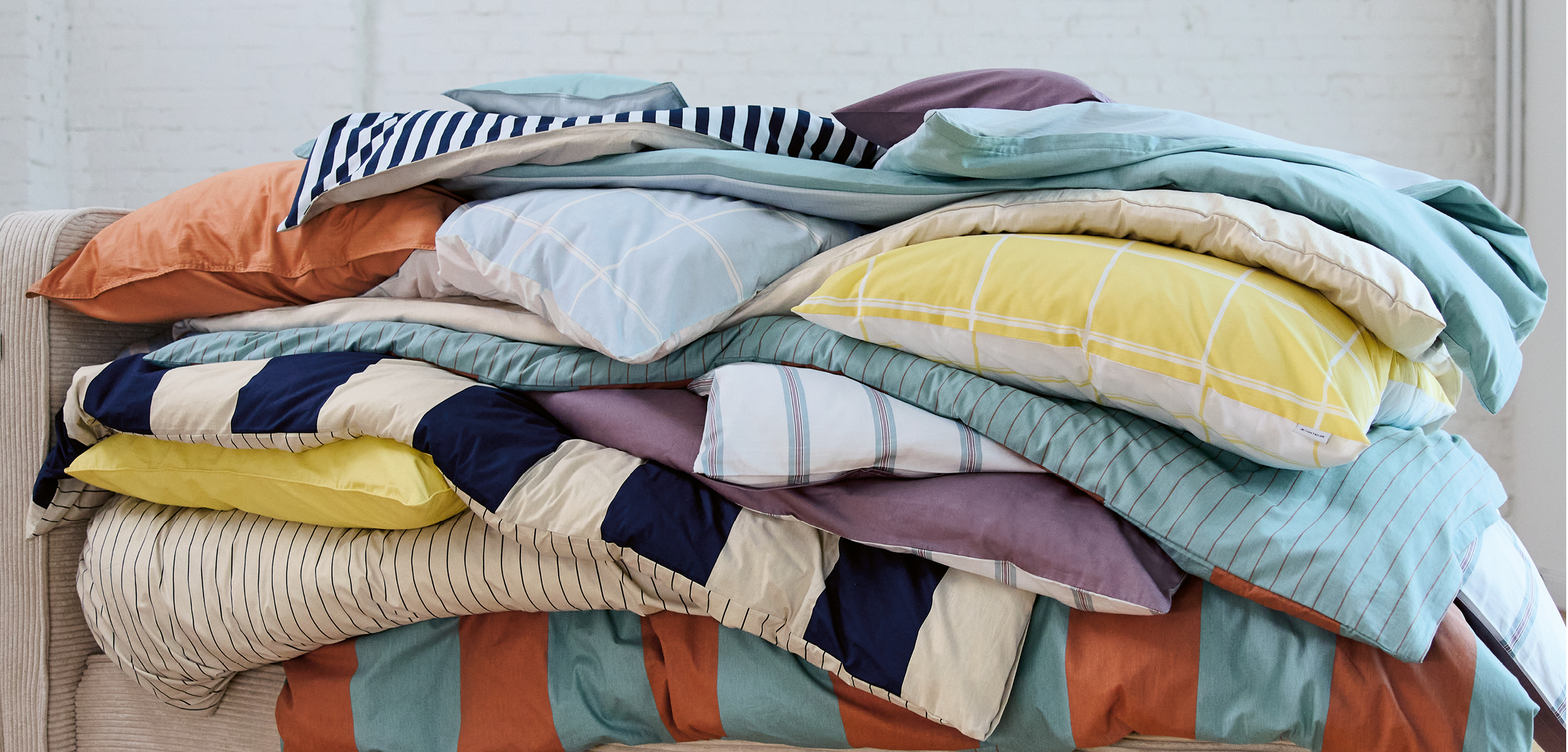 Ein farbenfroher Stapel Sommerbettwäsche mit verschiedenen Designs: Streifen, Karos, großflächige Blockstreifen und unifarbene Varianten in Blau, Türkis, Gelb, Orange, Beige und Violett.