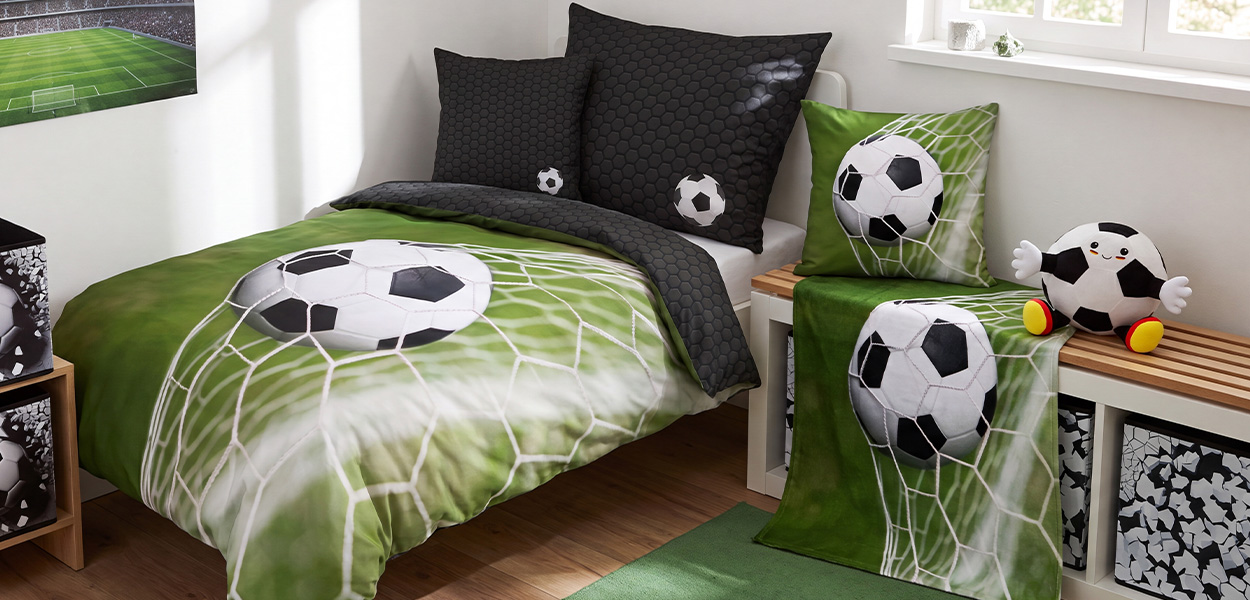Zu sehen ist ein helles Kinderzimmer im Fußball-Look. Bettwäsche, Kissen und Vorhang sind in Grün, Weiß und Schwarz gestaltet und zeigen jeweils einen Fußball im Tornetz. Dazu passen die schwarzen Kissen mit Wabenmuster und kleinen Fußballmotiven sowie die Aufbewahrungsboxen. Als dekorativer Blickfang sitzt ein Fußball-Kuscheltier auf der Bank.