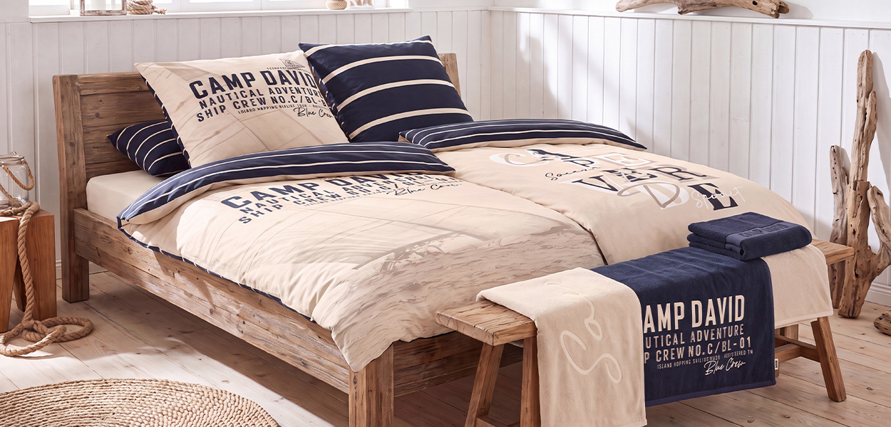 Das Bild zeigt ein gemütlich eingerichtetes Schlafzimmer im maritimen, rustikalen Stil. Im Mittelpunkt steht ein breites Holzbett mit Bettwäsche von **Camp David**. Die Farben sind überwiegend **Creme, Beige und Dunkelblau**, ergänzt durch navy-weiß gestreifte Kissen und Bettdeckenränder. Auf der Bettwäsche befinden sich markante Schriftzüge und nautisch inspirierte Grafiken. Als Dekoration liegen am Fußende gefaltete Handtücher bzw. eine Decke in Beige und Dunkelblau. Der helle Raum mit weißer Holzvertäfelung, Treibholz, Seilen und einem geflochtenen Teppich unterstreicht den entspannten Küsten- bzw. Strandhaus-Look.