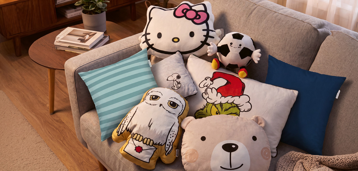 Kissen: Verschiedene Motivkissen mit Hello Kitty, Snoopy, Grinch, Hedwig, Fußball und Bär auf einem grauen Sofa.