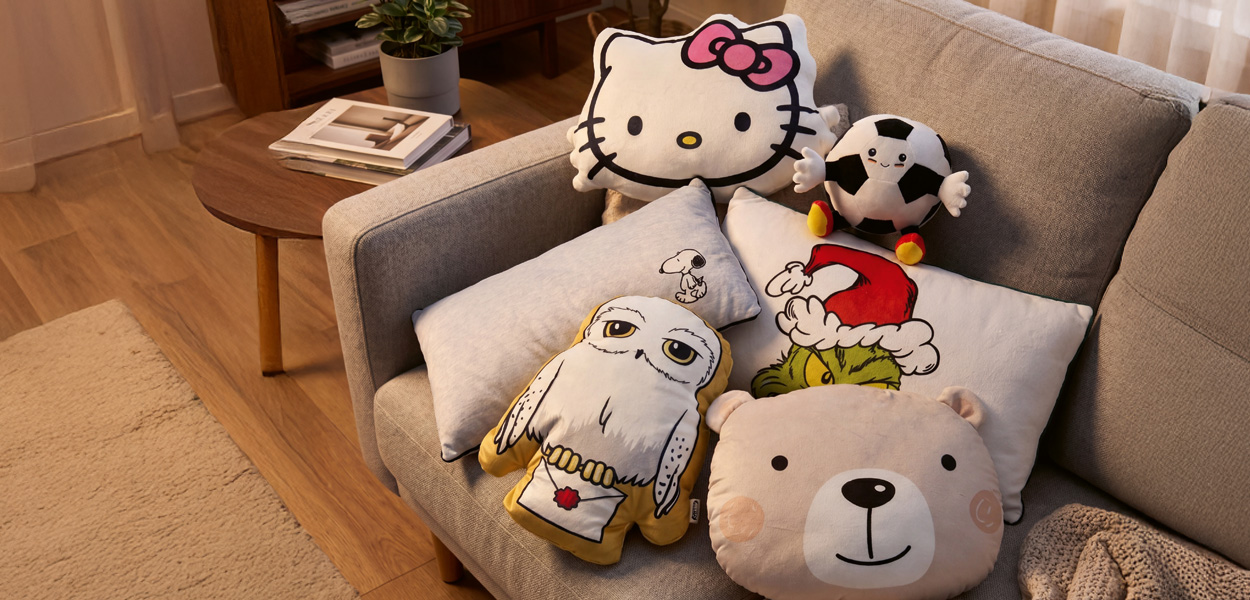 Kissen: Verschiedene Motivkissen auf einem grauen Sofa, darunter Hello Kitty, Snoopy, Grinch und ein Fußball.