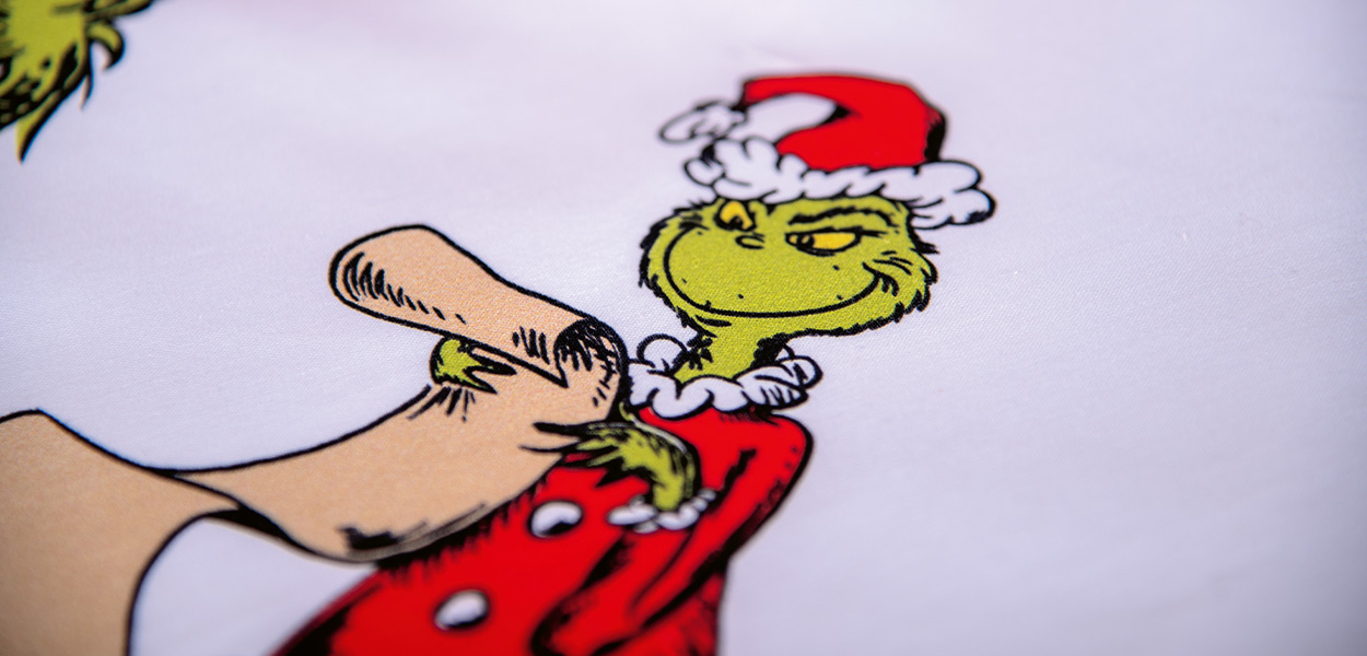Das Bild zeigt eine Nahaufnahme von Winterbettwäsche mit dem Grinch im Weihnachtsmann-Outfit. Er trägt eine rote Zipfelmütze und einen roten Mantel, während eine Hand seine Schulter berührt. Das Motiv wirkt weihnachtlich, humorvoll und passend zur Wintersaison.