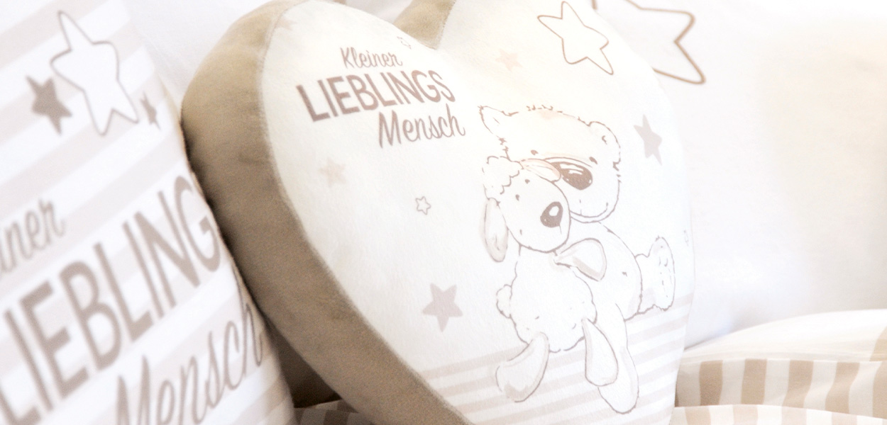 Kissen: Nahaufnahme eines beigen Dekokissens mit Snoopy-Motiv und dem Schriftzug „Kleiner Lieblingsmensch“ auf einem Bett.