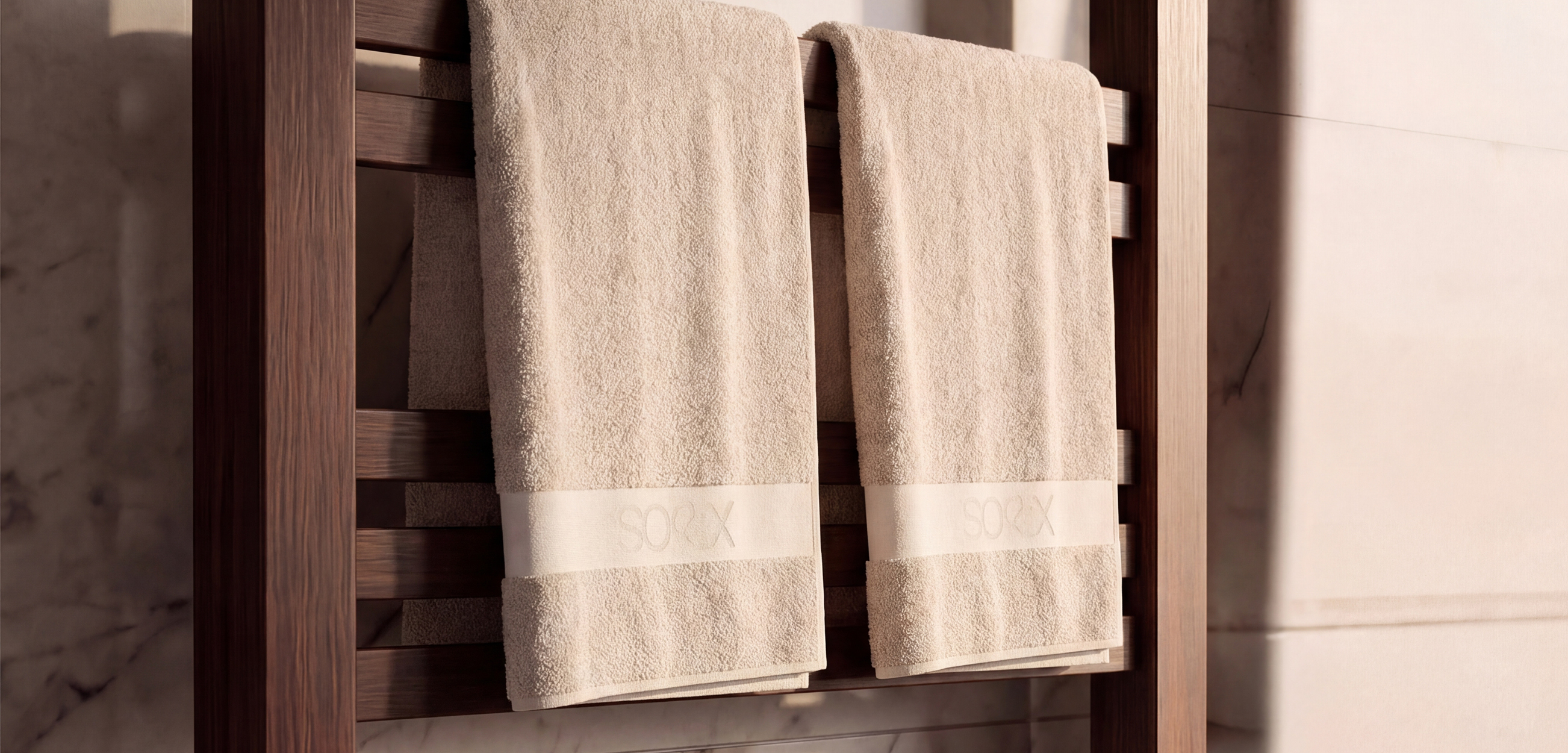 Zu sehen sind zwei hochwertige, beigefarbene Duschtücher auf einer dunklen Holz-Handtuchleiter in einem modernen Badezimmer. Die weiche Frottierstruktur wirkt besonders saugfähig und angenehm. Als dezentes Design-Detail haben beide Tücher eine breite, helle Bordüre mit der eingewebten Aufschrift „SOXX“. Warme Lichtstimmung, Holz und Marmor vermitteln einen ruhigen, eleganten Spa-Charakter.