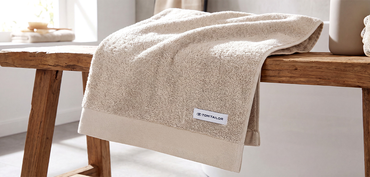 Zu sehen ist ein hochwertig wirkendes Duschtuch in einem hellen Beige-/Naturton. Es hat eine flauschige Frottierstruktur, eine breite glatte Bordüre und ein kleines, weißes Markenlabel mit der Aufschrift **„Tom Tailor“**.Das Tuch liegt locker über einer rustikalen Holzbank in einer hellen, wohnlichen Badezimmerumgebung.