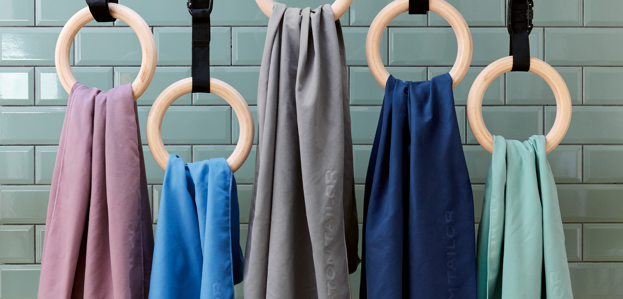 Das Bild zeigt das Fitnesstuch in fünf Farbvarianten: Rosé, Hellblau, Grau, Dunkelblau und Mint. Die Tücher hängen dekorativ über Holz-Turnringen vor einer türkisfarben gefliesten Wand. Auf den Tüchern ist jeweils ein dezentes, eingeprägtes „TOM TAILOR“-Logo zu sehen.
