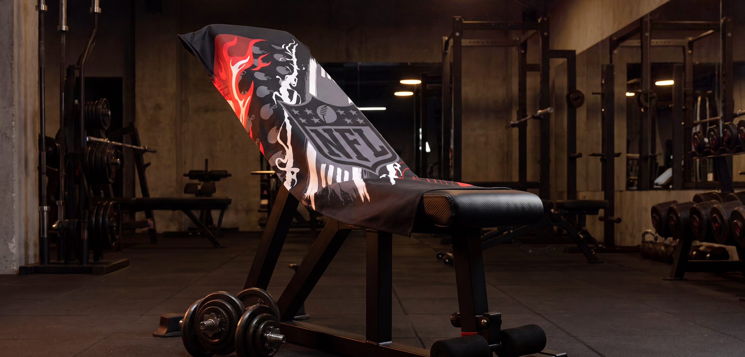 Das Bild zeigt ein Fitnesstuch im NFL-Design, das über die Rückenlehne einer Trainingsbank im Fitnessstudio gelegt ist. Das schwarze Tuch ist mit roten, weißen und grauen Football-Motiven sowie einem großen NFL-Logo gestaltet. Im Vordergrund liegen Hanteln, im Hintergrund sind weitere Fitnessgeräte zu sehen.