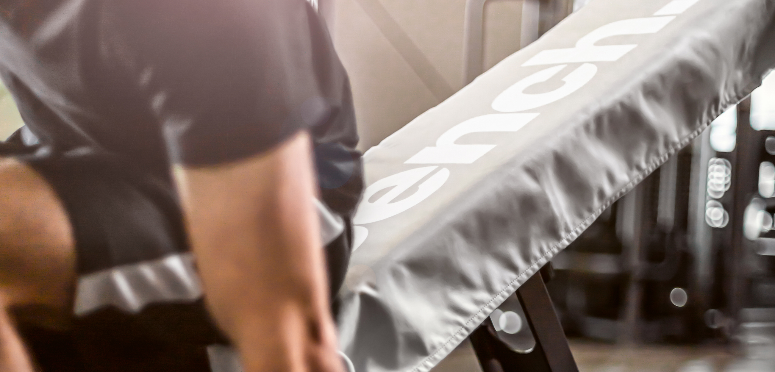 Das Bild zeigt ein graues **Fitnesstuch bzw. Sporthandtuch** mit großem weißem Schriftzug, das an einem Trainingsgerät liegt. Im Hintergrund ist eine Person beim Training zu sehen.