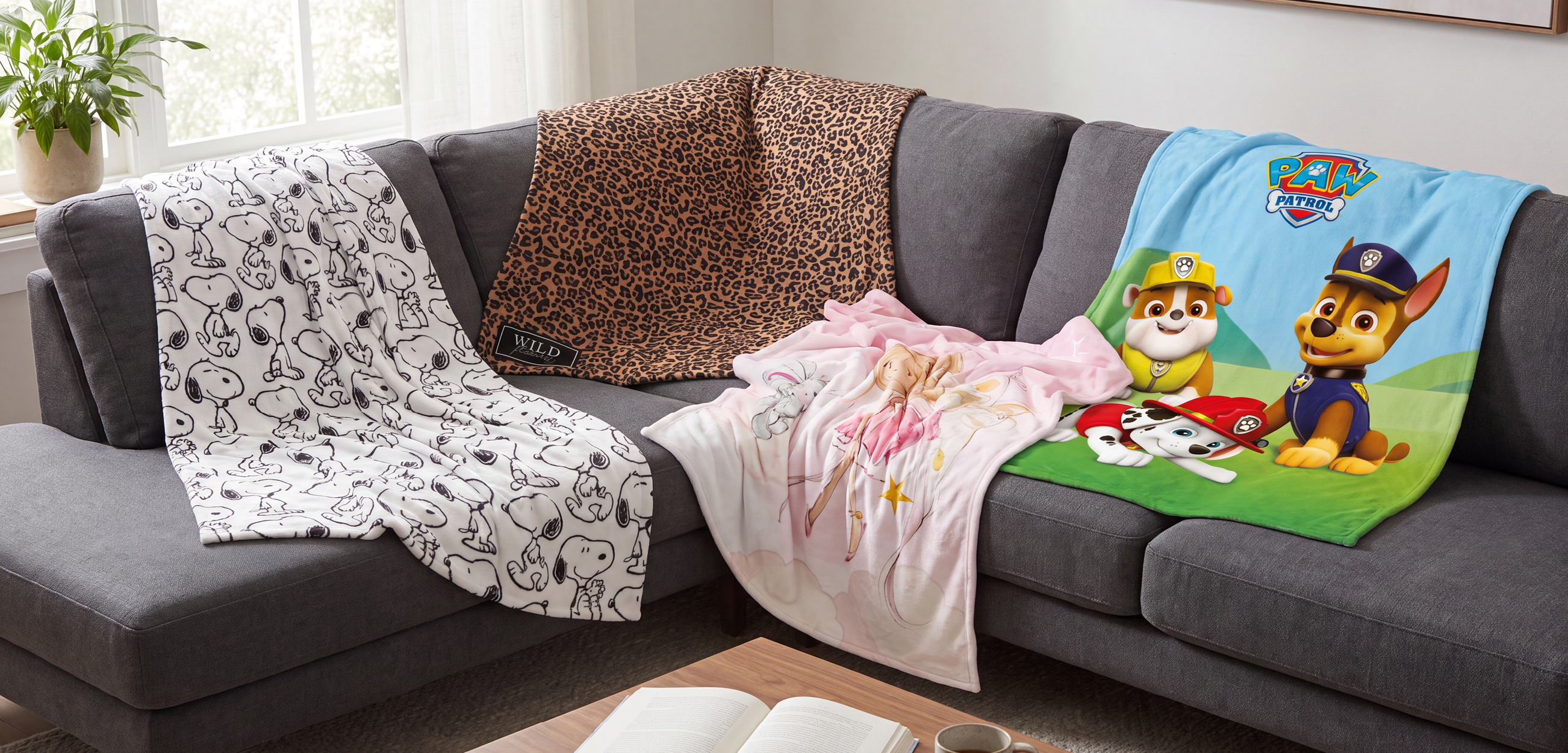 Das Bild zeigt eine Auswahl an Kuscheldecken in verschiedenen Designs auf einem Sofa: mit Snoopy-Motiv, Leopardenmuster, Prinzessinnenmotiv und PAW-Patrol-Design. Damit ist für Kinder und Erwachsene mit unterschiedlichen Lieblingsmotiven etwas dabei.