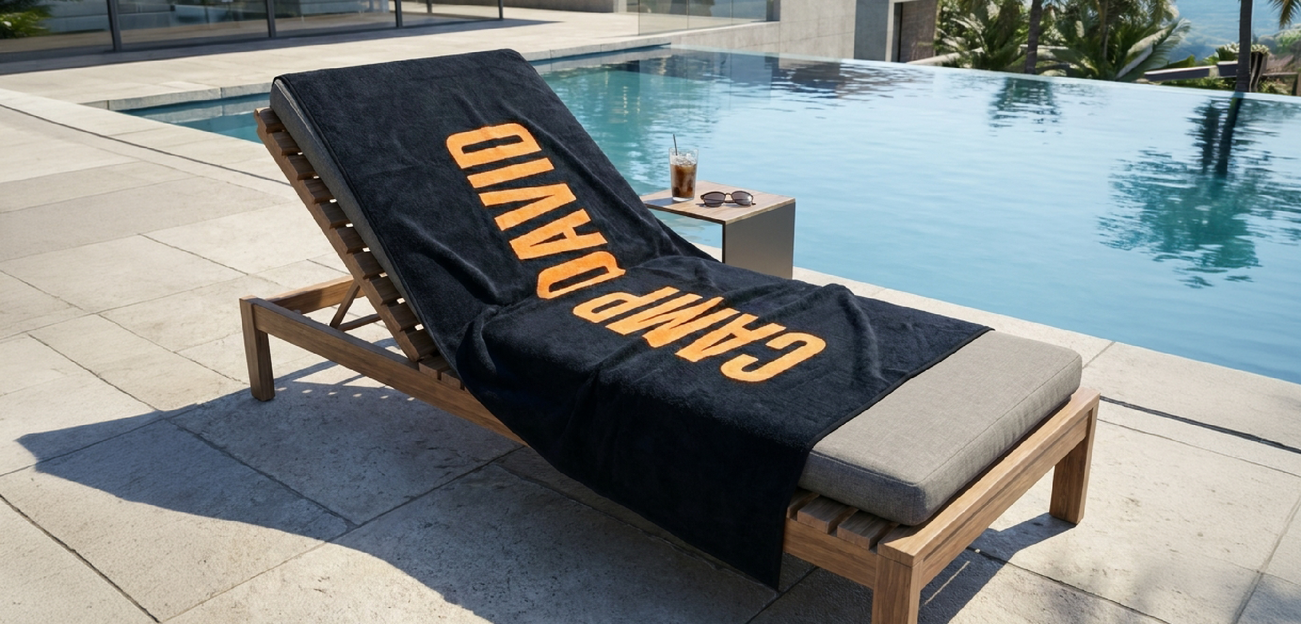 Dunkelblaue Handtücher im Strandtuch-Format mit großem orangefarbenem Schriftzug. Das weiche Badetuch liegt dekorativ auf einer Holzliege am Pool und vermittelt einen sportlich-modernen Sommerlook.