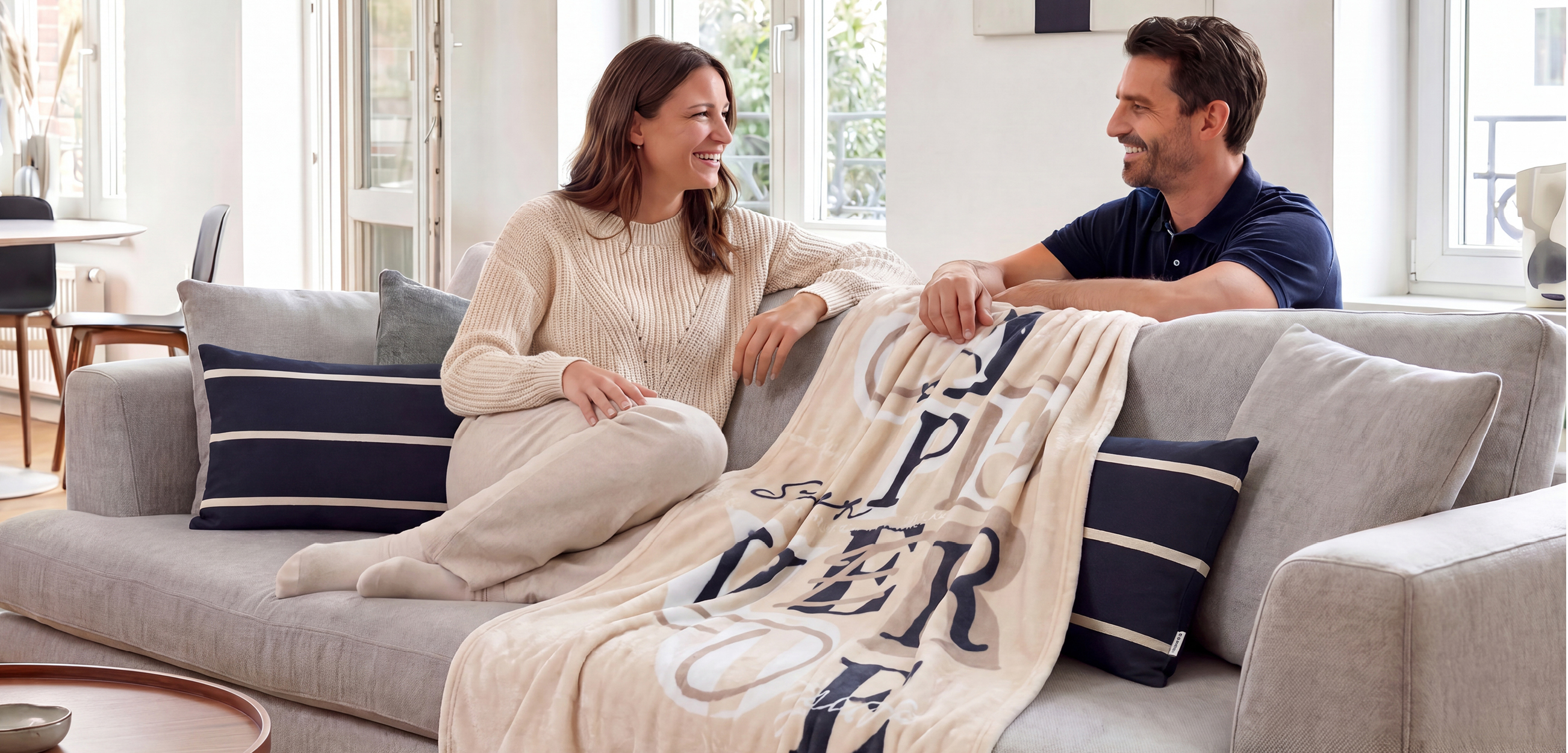 Das Bild zeigt ein helles, gemütliches Wohnzimmer. Ein Paar sitzt entspannt auf einem grauen Sofa. Auffällig sind die Heimtextilien: zwei dunkelblaue Kissen mit hellen Streifen sowie eine weiche, beigefarbene Kuscheldecke mit dunkelblauem, grafischem Muster. Die freundliche Einrichtung mit viel Tageslicht und warmen Naturtönen vermittelt Komfort und Wohlfühlatmosphäre.
