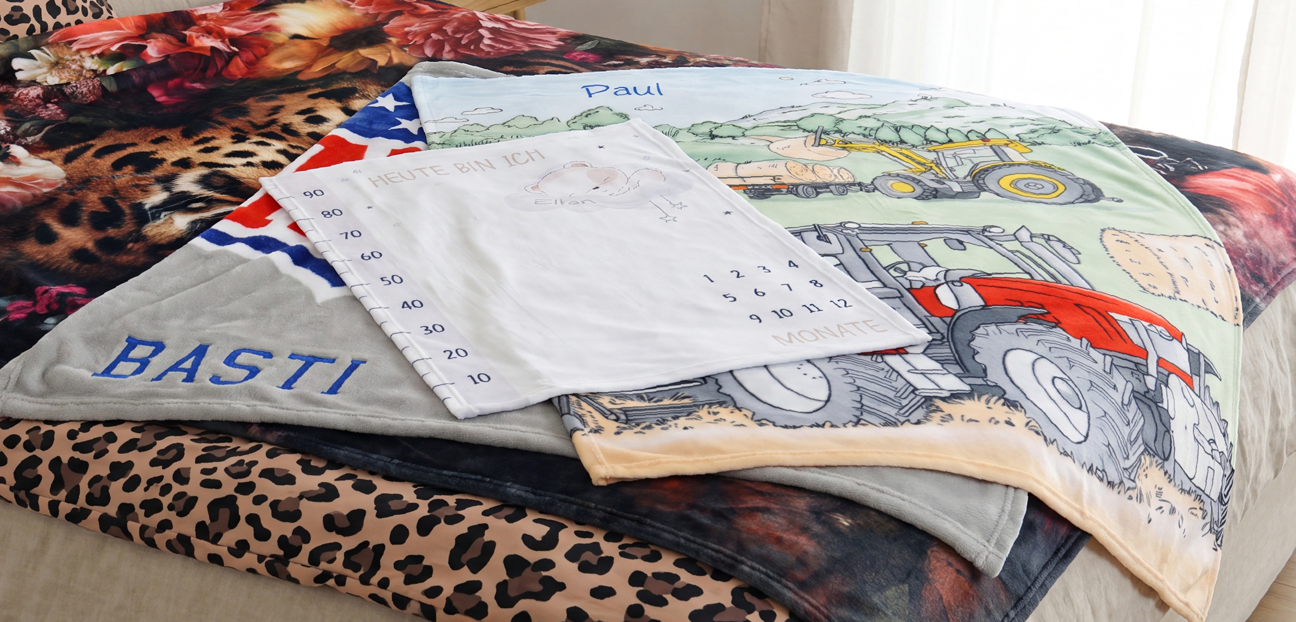Auf dem Bild sind mehrere kuschelige Decken mit unterschiedlichen Motiven und Personalisierungen zu sehen:- **Kinderdecke mit Traktor-Motiv** und dem Namen **„Paul“**
- **Graue Decke** mit dem gestickten Namen **„BASTI“**
- **Weiße Meilenstein-Decke für Babys** mit „Heute bin ich“, einem Bärenmotiv, Größen-Skala und Monatsangaben; sichtbar ist der Name **„Elian“**
- Weitere Decken mit **Leopardenmuster**, **Blumen-/Tiermotiv** und einer **USA-Flaggenoptik**Die Aufnahme zeigt gut, dass Namen, kindliche Motive und Erinnerungsdesigns individuell kombiniert werden können.