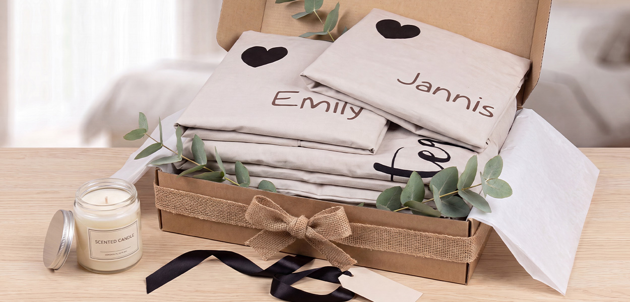 Das Bild zeigt einen liebevoll verpackten Geschenkkorb mit personalisierter Bettwäsche. Auf den hellen Kissenbezügen stehen die Namen **„Emily“** und **„Jannis“**, jeweils mit einem schwarzen Herz.Die Bettwäsche liegt in einer geöffneten Geschenkbox und ist mit Juteband, Schleife und Eukalyptus dekoriert. Daneben steht eine Duftkerze. Insgesamt wirkt es wie ein hochwertiges, persönliches Geschenk – zum Beispiel zur Hochzeit, zum Einzug oder für Paare.