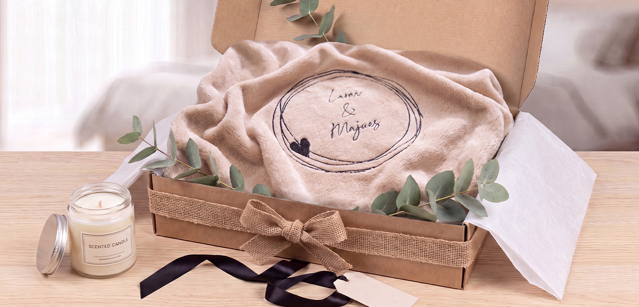 Das Bild zeigt eine **personalisierte, beigefarbene Kuscheldecke** in einer dekorativen Geschenkbox. Die Decke ist mit einem Namen bzw. einer Botschaft bestickt und wird zusammen mit Eukalyptuszweigen, Schleifenband und einer Duftkerze als hochwertiges Geschenk präsentiert.