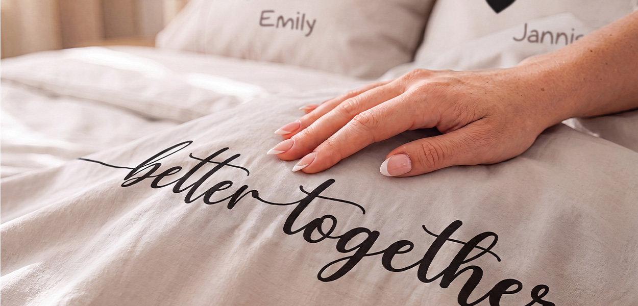 Das Bild zeigt eine helle, personalisierte Bettwäsche mit dem Schriftzug **„better together“**. Eine Hand streicht sanft über den Stoff. Im Hintergrund sind Kissen mit individuellen Namen wie **„Emily“** und **„Jannie“** zu sehen – die Szene vermittelt Nähe, Gemütlichkeit und eine persönliche Geschenkidee.