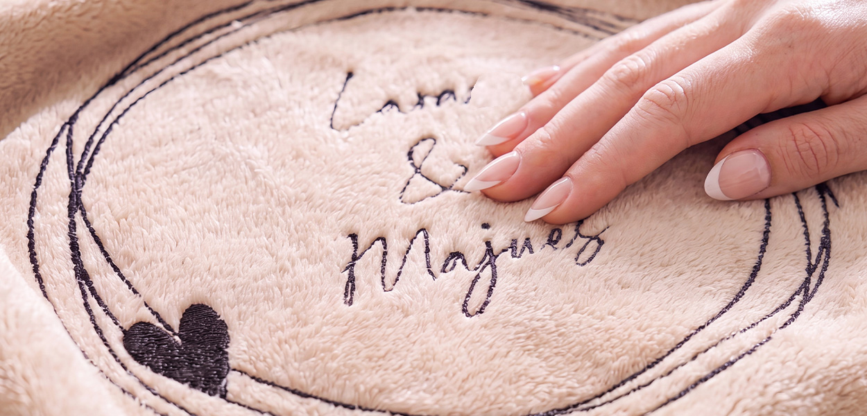 Zu sehen ist eine weiche, beigefarbene Kuscheldecke mit einer dunklen, gestickten Personalisierung. In einem kreisförmigen Motiv stehen die Namen „Lina & Magnus“, ergänzt durch ein kleines Herz. Eine Hand streicht über den flauschigen Stoff und betont die angenehme, hochwertige Haptik.