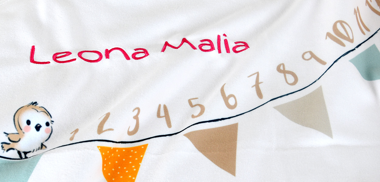 Weiße, personalisierbare Babydecke mit dem Namen „Leona Malia“ in pinker Stickschrift, Zahlenreihe und bunten Wimpeln.