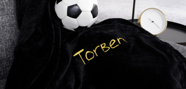 personalisierbare Heimtextilien: Schwarze, kuschelige Decke mit goldgelber Personalisierung „Torben“, dekoriert auf einem grauen Sofa neben einem Fußball.