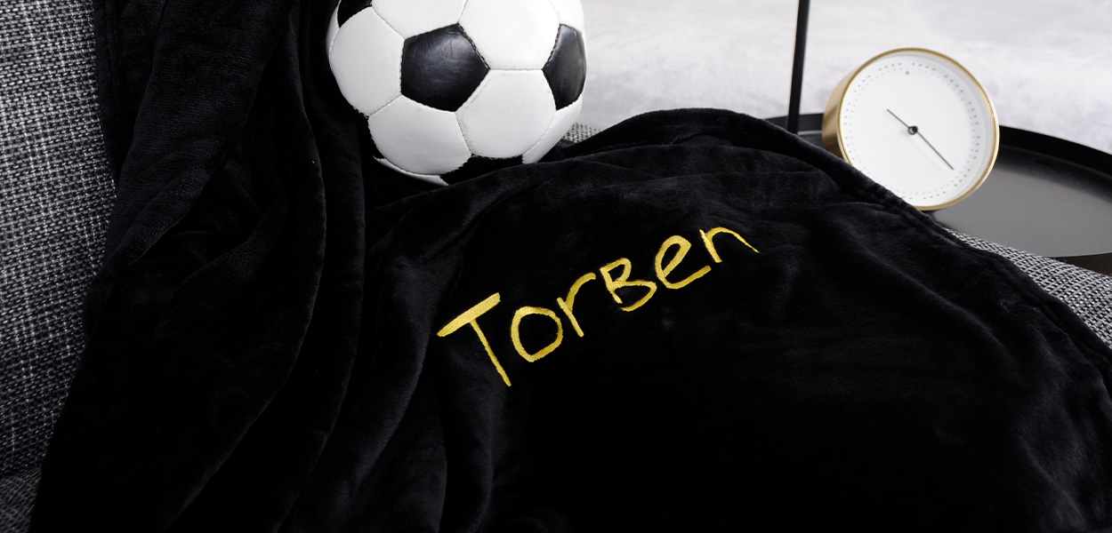 Schwarze, kuschelige Decke mit goldgelber Personalisierung „Torben“, dekoriert auf einem grauen Sofa neben einem Fußball.