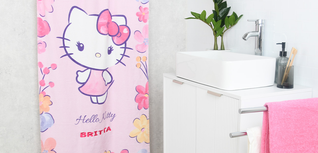 Ein rosa Hello-Kitty-Handtuch mit Blumenmuster und dem personalisierten Namen „BRITTA“. Es hängt in einem modernen Badezimmer neben dem Waschbecken.