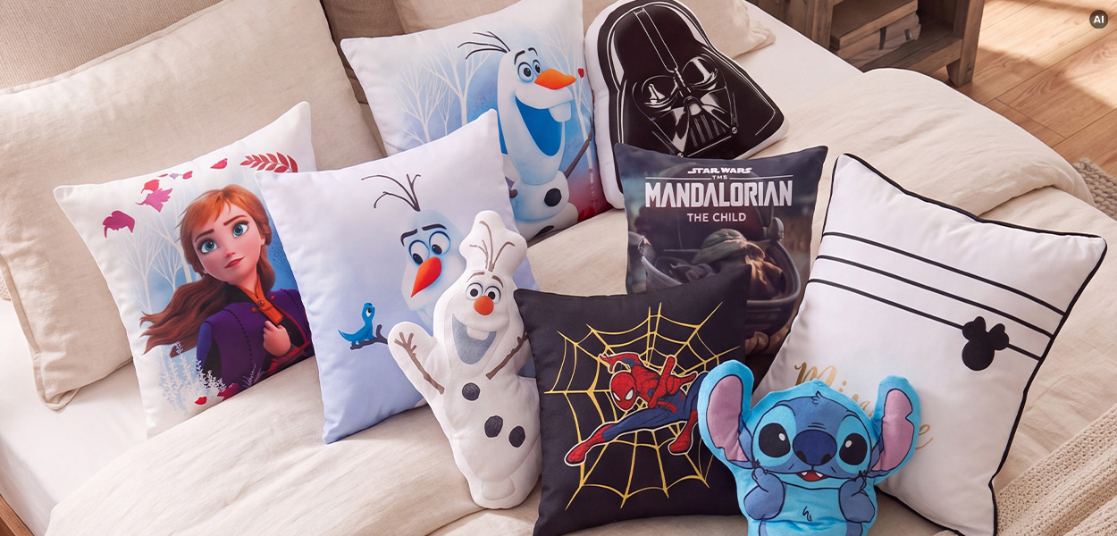 Disney Kissen: Auf dem Bild liegen verschiedene Disney-Kissen auf einem Bett. Zu sehen sind Motive von Anna und Olaf aus „Die Eiskönigin“, Darth Vader, The Mandalorian mit Grogu, Spider-Man, Stitch sowie ein weißes Kissen mit Mickey-Mouse-Silhouette.