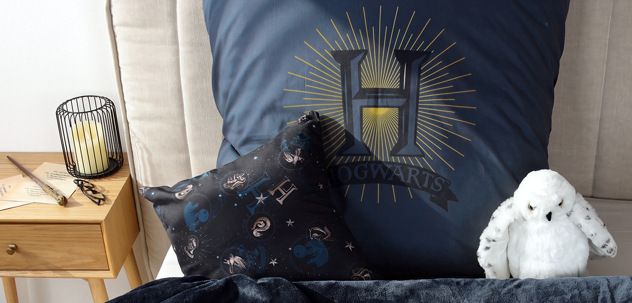 Harry Potter Heimtextilien: Harry-Potter-Bettwäsche in Dunkelblau mit großem Hogwarts-Logo, magischen Symbolen und passendem Dekokissen auf einem Bett.